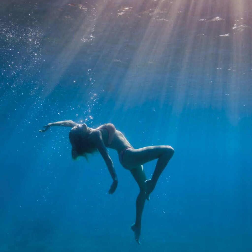 Woman floating underwater with sunlight beams representing the Indulge 120-minute massage session. Photograph by Kelly Hsiao.